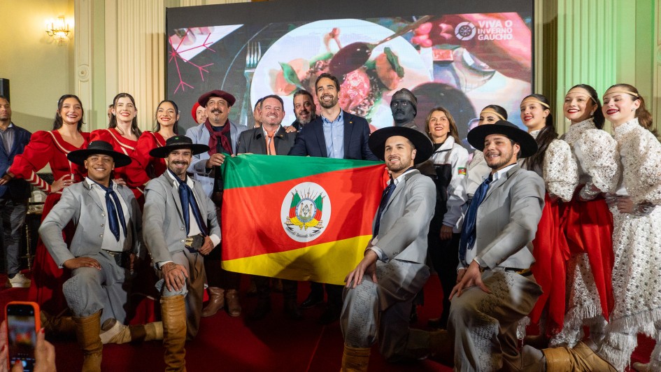 Foto posada com em torno de 20 homens e mulheres. Leite e Santini estão no cengtro, segurando a bandeira do RS. Ao redor, pessoas vestindo trajes tipicamente gaúchos. Todos sorriem.