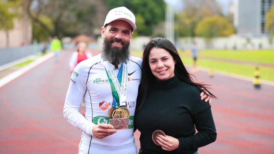 Foto posada em pista de corrida, ao ar livre, de Tiarles, com roupa de corrida, com várias medalhas penduradas no pescoço. Ao seu lado está Thalyne, de blusa preta. Ambos sorriem, numa tomada em plano americano frontal.