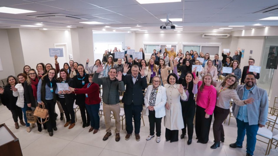 A imagem mostra um grupo grande de pessoas reunidas em um ambiente interno claro e moderno, possivelmente uma sala de eventos ou auditório. Elas estão posicionadas de frente para a câmera, muitas sorrindo e acenando com uma das mãos levantadas. Algumas seguram certificados, indicando que o momento é de celebração ou formatura. As cadeiras ao fundo estão parcialmente ocupadas, e há um projetor no teto. O grupo é diverso, composto majoritariamente por mulheres, e todos parecem estar vestidos de forma casual ou social.
