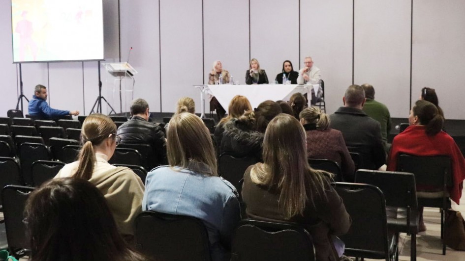 A imagem é de um ambiente formal de conferência ou seminário, com quatro palestrantes em um palco, uma plateia atenta e uma tela de apresentação ao lado.