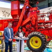 A foto mostra o Secretário da Cultura, Eduardo Loureiro, e a diretora do MEA, Karina Muniz Viana, em pé diante de uma mesa redonda, formalizando o acordo. Ao lado deles, uma grande máquina agrícola vermelha, antiga, está em exibição. Ao fundo, painéis com ilustrações sobre a evolução agrícola decoram a parede.
