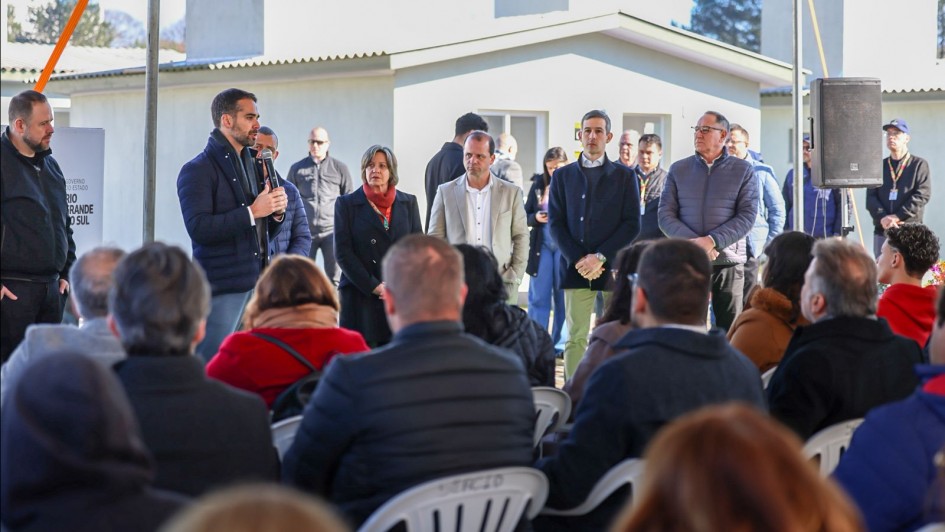 A foto mostra o governador Eduardo Leite no centro, discursando em um microfone. Ele está em um palco ao ar livre, vestindo um casaco azul-escuro. Atrás dele, há uma plateia sentada em fileiras, e ao fundo, é possível ver um grupo de casas brancas, recém-construídas.