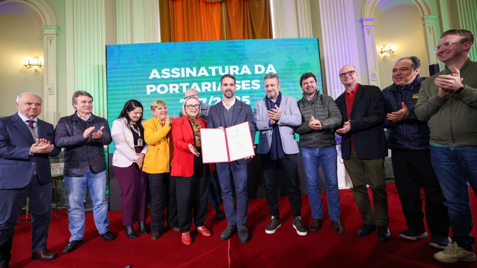 A imagem mostra um grupo de 11 pessoas, em pé, em um palco com um tapete vermelho. No centro, dois homens estão segurando um documento. Um deles está de terno, o outro está de blazer. Atrás deles, há um grande painel digital com o texto "ASSINATURA DA PORTARIA SES". As outras pessoas estão aplaudindo, algumas olhando para o documento, outras para o público. O cenário inclui cortinas de veludo e paredes decoradas.