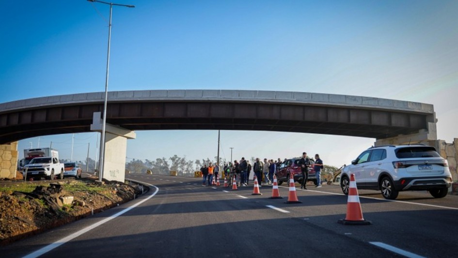 A imagem mostra um grupo de pessoas em pé em uma rodovia, próximo a um viaduto. O viaduto atravessa a rodovia na parte superior da imagem. Algumas pessoas parecem estar em uma cerimônia ou evento de inauguração. Há cones de trânsito laranja e carros estacionados, incluindo um SUV branco à direita e uma van branca ao fundo. O céu está limpo e azul.