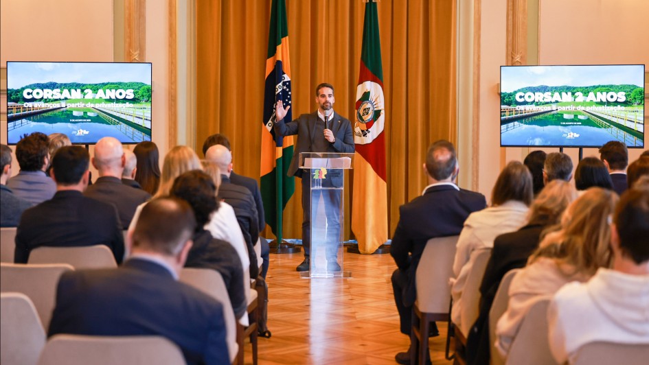 O governador do Rio Grande do Sul, Eduardo Leite, está em pé em um púlpito, falando para uma plateia sentada. Ele gesticula com a mão direita enquanto a esquerda está sobre o púlpito. Atrás dele, há duas bandeiras: a do Brasil e a do Rio Grande do Sul. Em cada lado do palco, duas telas de LED exibem uma imagem de uma ponte sobre a água, com o logo "CORSAN 2 ANOS" no canto superior esquerdo.