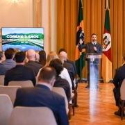 O governador do Rio Grande do Sul, Eduardo Leite, está em pé em um púlpito, falando para uma plateia sentada. Ele gesticula com a mão direita enquanto a esquerda está sobre o púlpito. Atrás dele, há duas bandeiras: a do Brasil e a do Rio Grande do Sul. Em cada lado do palco, duas telas de LED exibem uma imagem de uma ponte sobre a água, com o logo "CORSAN 2 ANOS" no canto superior esquerdo.