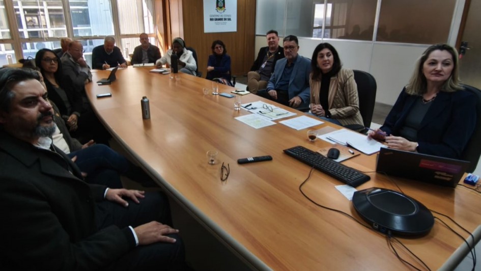 A foto mostra diversas pessoas sentadas ao redor de uma mesa de reunião. Em cima da mesa, há papéis, óculos, canetas e notebooks.