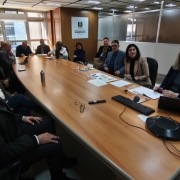 A foto mostra diversas pessoas sentadas ao redor de uma mesa de reunião. Em cima da mesa, há papéis, óculos, canetas e notebooks. 