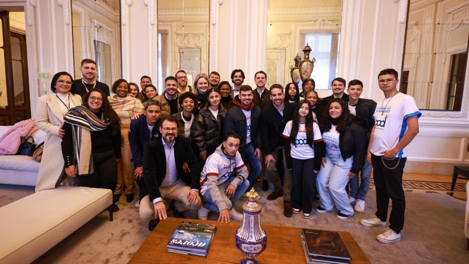 Grupo de pessoas posando em sala elegante com espelhos ornamentados, móveis clássicos e mesa com livros e vaso decorativo. Alguns usam camisetas iguais com texto. Ambiente sofisticado e acolhedor.