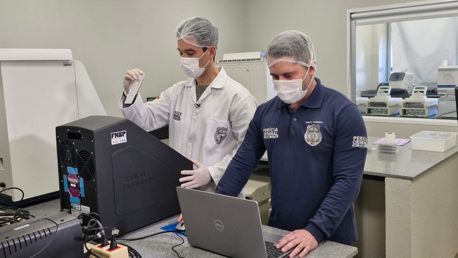 A imagem mostra dois homens, ambos de touca e máscara de proteção, em um laboratório. O homem da esquerda veste um jaleco branco e luvas, e segura um pequeno tubo de ensaio. O homem da direita veste uma camiseta azul escura com a inscrição "PERÍCIA CRIMINAL" e o símbolo da Polícia Federal. Ele está sentado em frente a um notebook Dell, com a mão direita sobre o mouse e a esquerda sobre o teclado, aparentemente interagindo com o computador.

Entre os dois, há um equipamento grande e preto com o logo "Applied Biosystems" e a sigla "FNSP" visível. O ambiente de fundo é um laboratório, com bancadas cinzas de granito, diversos outros equipamentos de laboratório e janelas com molduras brancas que separam o espaço de outro cômodo. O homem de jaleco tem cabelos escuros, e o de camiseta azul tem barba.