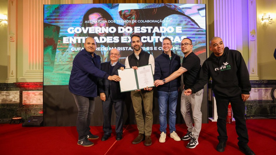 A foto mostra um grupo de seis homens, com o governador Eduardo Leite no centro, posando para a câmera em um palco. Eles estão segurando um grande certificado ou documento de cor verde. O ato ocorreu no Palácio Piratini.