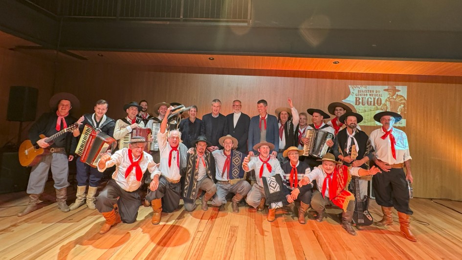 Grupo de homens posando para foto em um palco de madeira, com parede de fundo clara. A maioria está vestida com trajes típicos gaúchos — bombacha, camisa branca, colete ou poncho, lenço vermelho no pescoço e chapéu. Alguns seguram instrumentos musicais, como acordeons, violão e baixo. Ao fundo, três homens de terno preto estão no centro da imagem. No canto direito, há um telão com a imagem e o texto “Registro do Gênero Musical Bugio”. A iluminação é quente, destacando as cores vivas das roupas e o brilho do piso de madeira.