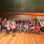 Grupo de homens posando para foto em um palco de madeira, com parede de fundo clara. A maioria está vestida com trajes típicos gaúchos — bombacha, camisa branca, colete ou poncho, lenço vermelho no pescoço e chapéu. Alguns seguram instrumentos musicais, como acordeons, violão e baixo. Ao fundo, três homens de terno preto estão no centro da imagem. No canto direito, há um telão com a imagem e o texto “Registro do Gênero Musical Bugio”. A iluminação é quente, destacando as cores vivas das roupas e o brilho do piso de madeira.