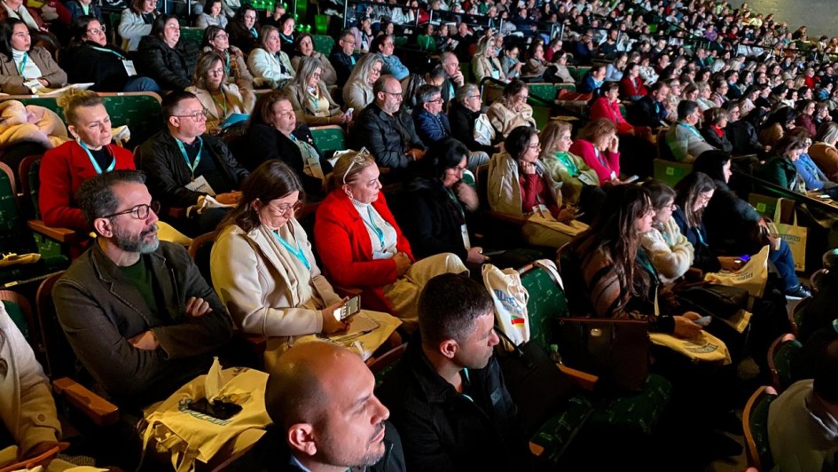 Auditório cheio, com muitas pessoas sentadas em cadeiras verdes, assistindo a um evento iluminado.