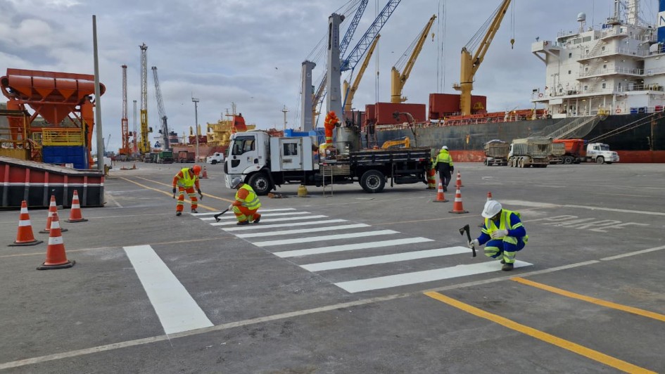 Descrição para acessibilidade: Trabalhadores com coletes refletivos e capacetes realizam a pintura de uma faixa de pedestres em área portuária. Ao fundo, há um caminhão de apoio, cones de sinalização, guindastes e grandes navios de carga atracados, com contêineres e equipamentos de operação portuária visíveis. O céu está nublado.