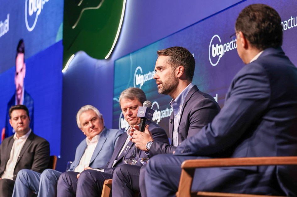 A imagem retrata um cenário de painel de discussão, com cinco pessoas sentadas no palco. Ao fundo, há uma tela grande exibindo repetidamente o logotipo "btg pactual". Um dos participantes, o governador Eduardo Leite está falando ao microfone, enquanto os demais escutam com atenção.