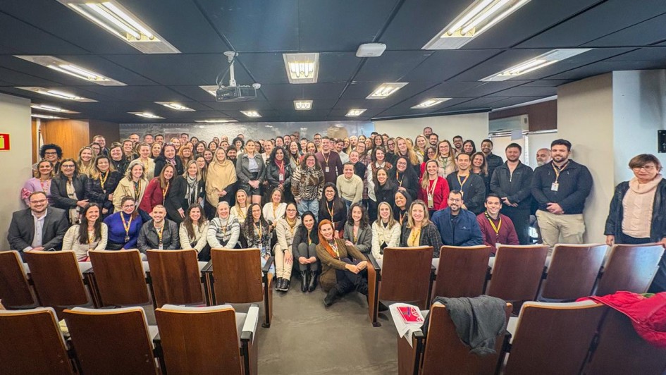 Fotografia de um grande grupo de pessoas reunidas em um auditório. Elas estão dispostas em várias fileiras, com algumas sentadas nas cadeiras da frente e a maioria em pé ao fundo. O ambiente é iluminado por luzes no teto e possui cadeiras de madeira com estofado claro. No centro, há uma pessoa posicionada levemente à frente, cercada pelos demais participantes. Ao fundo, vê-se uma parede com revestimento claro e um projetor suspenso no teto. Todos posam sorrindo para a foto, transmitindo clima de confraternização ou evento corporativo.