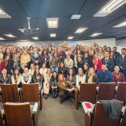 Fotografia de um grande grupo de pessoas reunidas em um auditório. Elas estão dispostas em várias fileiras, com algumas sentadas nas cadeiras da frente e a maioria em pé ao fundo. O ambiente é iluminado por luzes no teto e possui cadeiras de madeira com estofado claro. No centro, há uma pessoa posicionada levemente à frente, cercada pelos demais participantes. Ao fundo, vê-se uma parede com revestimento claro e um projetor suspenso no teto. Todos posam sorrindo para a foto, transmitindo clima de confraternização ou evento corporativo.