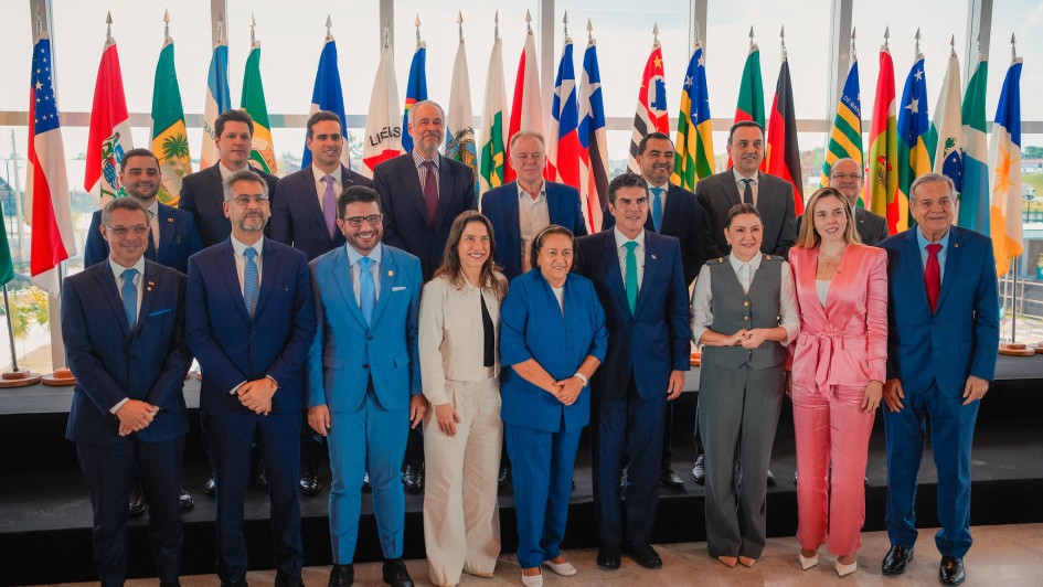 A imagem mostra um grupo de cerca de 20 pessoas, homens e mulheres, de pé em três fileiras. Eles estão vestidos com roupas sociais, como ternos e tailleurs. A maioria dos homens veste ternos em tons de azul ou cinza escuro, enquanto as mulheres usam tailleurs em cores como branco, azul claro e rosa. A pessoa em destaque no centro da primeira fila é um homem alto com cabelo escuro, vestindo um terno azul-celeste. Ele está sorrindo. Atrás do grupo, há uma fileira de mastros com bandeiras de diferentes estados brasileiros e a bandeira do Brasil. O ambiente parece ser um espaço de reunião ou evento oficial, com iluminação clara.