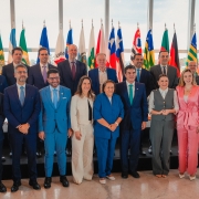A imagem mostra um grupo de cerca de 20 pessoas, homens e mulheres, de pé em três fileiras. Eles estão vestidos com roupas sociais, como ternos e tailleurs. A maioria dos homens veste ternos em tons de azul ou cinza escuro, enquanto as mulheres usam tailleurs em cores como branco, azul claro e rosa. A pessoa em destaque no centro da primeira fila é um homem alto com cabelo escuro, vestindo um terno azul-celeste. Ele está sorrindo. Atrás do grupo, há uma fileira de mastros com bandeiras de diferentes estados brasileiros e a bandeira do Brasil. O ambiente parece ser um espaço de reunião ou evento oficial, com iluminação clara.