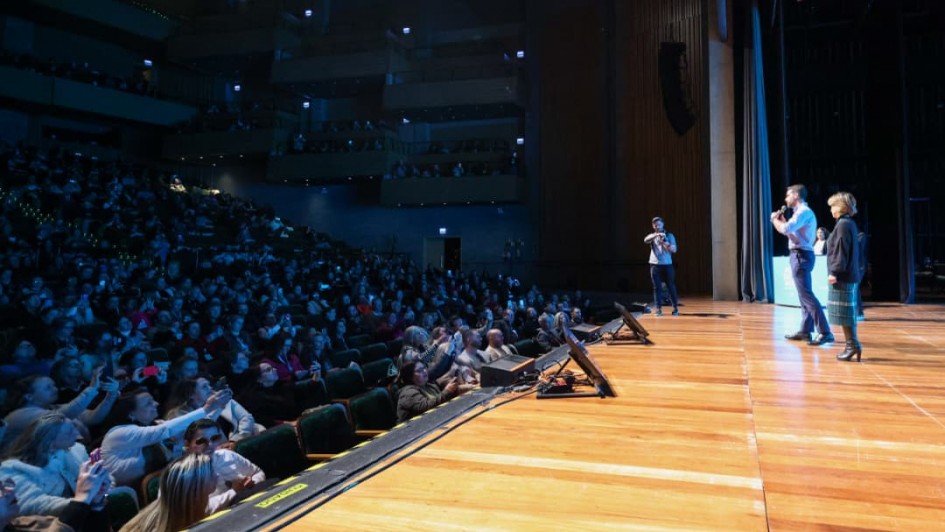 Auditório lotado com várias pessoas assistindo a uma apresentação. Três pessoas estão no palco de madeira, uma fala ao microfone. Plateia atenta, alguns registros com celular. Ambiente amplo e com vários níveis de assentos.