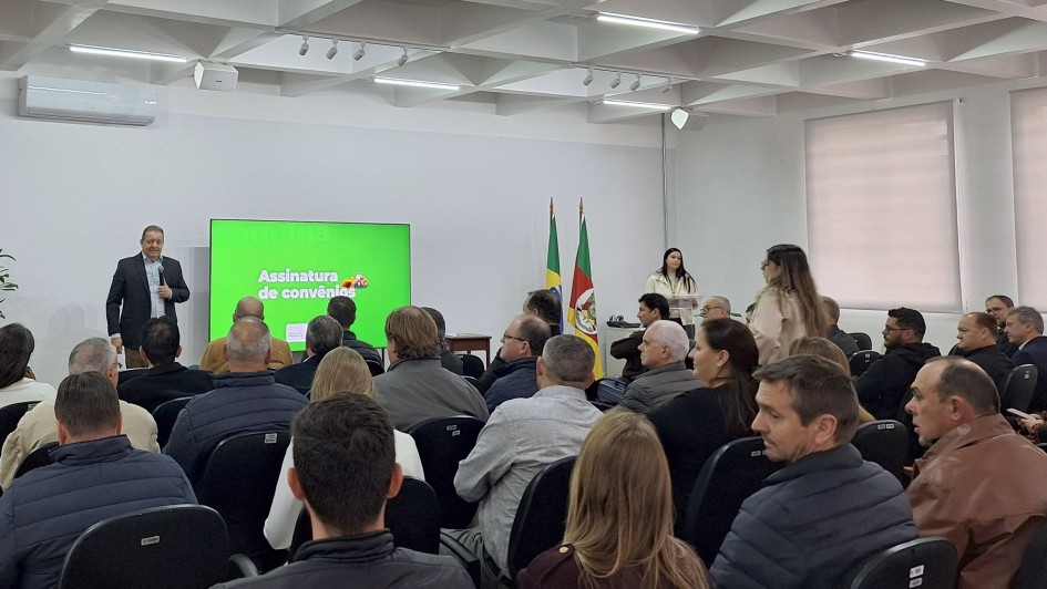 Esta é uma fotografia de um evento formal em um auditório, visto da parte de trás da plateia, olhando em direção ao palco.

No canto superior esquerdo, um homem de terno escuro está em pé ao lado de um telão verde-claro. O telão exibe o texto "Assinatura de convênios". O homem gesticula com as mãos enquanto fala em um microfone que está em um pedestal.

A plateia, composta por dezenas de pessoas, está sentada em cadeiras escuras, voltada para o palco. No lado direito do palco, há duas bandeiras de mastros altos, uma do Brasil e outra do Rio Grande do Sul.

As paredes da sala são de cor clara, e o teto é baixo, com luminárias embutidas. A iluminação é uniforme. O ambiente parece ser um evento de assinatura de documentos ou uma reunião oficial.