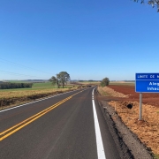 Imagem de uma estrada asfaltada, com faixas centrais amarelas e laterais brancas, que se estende em direção ao horizonte em meio a uma paisagem rural. À direita, há uma placa azul indicando o limite entre os municípios de Alegria e Inhacorá. O céu está limpo e azul, e há campos cultivados dos dois lados da via.