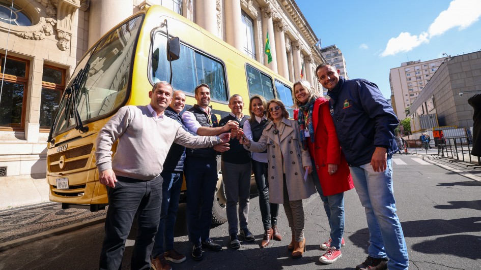 Esta é uma fotografia de um grupo de oito pessoas, seis homens e duas mulheres, em pé na rua, posando para a câmera em frente a um grande ônibus escolar amarelo.

O grupo está disposto em um semicírculo. Todos sorriem e estendem as mãos em direção ao centro, como em um aperto de mão coletivo. O homem no centro, com barba, veste um terno azul-marinho. A mulher à sua direita veste um casaco bege de estilo trench coat.

O ônibus amarelo, com a marca "IVECO", domina o lado esquerdo da imagem. Ao fundo, há um grande prédio com arquitetura formal, colunas e janelas altas. O céu é azul com nuvens brancas. O ambiente é de celebração e oficial.