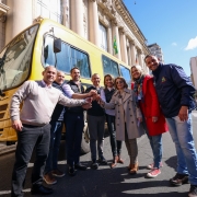 Esta é uma fotografia de um grupo de oito pessoas, seis homens e duas mulheres, em pé na rua, posando para a câmera em frente a um grande ônibus escolar amarelo.

O grupo está disposto em um semicírculo. Todos sorriem e estendem as mãos em direção ao centro, como em um aperto de mão coletivo. O homem no centro, com barba, veste um terno azul-marinho. A mulher à sua direita veste um casaco bege de estilo trench coat.

O ônibus amarelo, com a marca "IVECO", domina o lado esquerdo da imagem. Ao fundo, há um grande prédio com arquitetura formal, colunas e janelas altas. O céu é azul com nuvens brancas. O ambiente é de celebração e oficial.