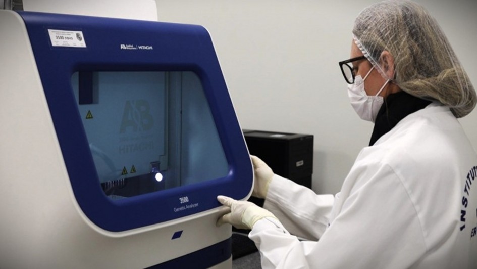 A foto mostra uma mulher de perfil, em um laboratório. Ela usa um jaleco branco, óculos, luvas e uma máscara de proteção. A mulher está em frente a uma máquina de cor azul e branca. A máquina parece ser um analisador genético. A mulher está tocando no painel frontal da máquina, com uma expressão focada.