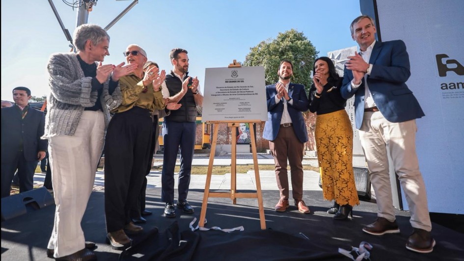 A foto mostra o governador Eduardo Leite e um grupo de pessoas batendo palmas em frente a um totem de madeira com uma placa de inauguração. A placa tem a inscrição "GOVERNO DO ESTADO DO RIO GRANDE DO SUL" e os logos de algumas secretarias. O ato acontece ao ar livre, com a luz do sol, e ao fundo, há um prédio de tijolos à vista.