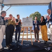 A foto mostra o governador Eduardo Leite e um grupo de pessoas batendo palmas em frente a um totem de madeira com uma placa de inauguração. A placa tem a inscrição "GOVERNO DO ESTADO DO RIO GRANDE DO SUL" e os logos de algumas secretarias. O ato acontece ao ar livre, com a luz do sol, e ao fundo, há um prédio de tijolos à vista.