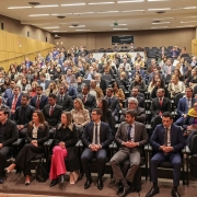 Esta é uma fotografia de um grande auditório em perspectiva, mostrando a plateia sentada e vista do lado do palco.

A plateia, composta por centenas de pessoas, está sentada em cadeiras pretas em filas inclinadas. A maioria dos participantes é adulta e está vestida com trajes formais, como ternos e blazers. Algumas pessoas usam camisas sociais, e há várias mulheres vestindo blazers e camisas sociais.

As paredes laterais do auditório são revestidas com painéis acústicos de cor amarela. O teto é alto e branco, com iluminação embutida. O ambiente é formal e a foto parece ter sido tirada durante um evento, como uma palestra ou uma cerimônia de posse.