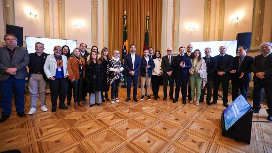 Grupo de pessoas em pé numa sala elegante, com chão de madeira, paredes ornamentadas, bandeiras ao fundo e telas grandes dos dois lados. Iluminação com lustres.