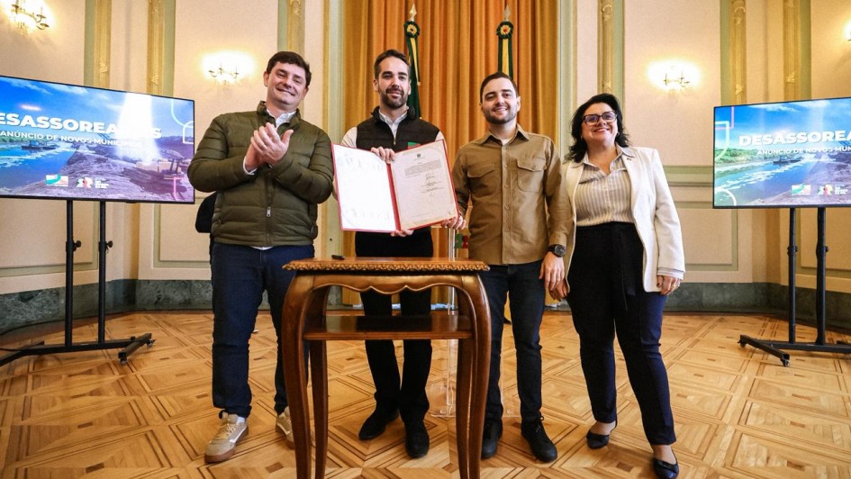 A fotografia mostra quatro pessoas, dois homens e duas mulheres, em pé, posando para a câmera em um ambiente formal. O governador Eduardo Leite está no centro, segurando um documento aberto com uma capa vermelha. Ele veste uma camisa preta e um colete preto, e tem barba. Ao seu lado esquerdo, o secretário Marcelo Caumo, um homem de jaqueta verde oliva aplaude. À direita de Leite, o vice-governador Gabriel Souza, um homem de camisa marrom, e a secretária Marjorie Kauffmann, uma mulher de casaco bege com óculos sorriem. Eles estão de pé sobre um piso de madeira com padrões geométricos. Atrás deles, há duas telas de LED, cada uma exibindo o logo do programa "DESASSOREAR RS" e o texto "ANÚNCIO DE NOVOS 67 MUNICÍPIOS". Também é possível ver as bandeiras do Brasil e do Rio Grande do Sul e cortinas douradas.