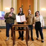 A fotografia mostra quatro pessoas, dois homens e duas mulheres, em pé, posando para a câmera em um ambiente formal. O governador Eduardo Leite está no centro, segurando um documento aberto com uma capa vermelha. Ele veste uma camisa preta e um colete preto, e tem barba. Ao seu lado esquerdo, o secretário Marcelo Caumo, um homem de jaqueta verde oliva aplaude. À direita de Leite, o vice-governador Gabriel Souza, um homem de camisa marrom, e a secretária Marjorie Kauffmann, uma mulher de casaco bege com óculos sorriem. Eles estão de pé sobre um piso de madeira com padrões geométricos. Atrás deles, há duas telas de LED, cada uma exibindo o logo do programa "DESASSOREAR RS" e o texto "ANÚNCIO DE NOVOS 67 MUNICÍPIOS". Também é possível ver as bandeiras do Brasil e do Rio Grande do Sul e cortinas douradas.