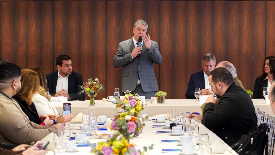 Esta é uma fotografia de um evento de negócios ou coletiva de imprensa, com um homem em pé discursando para um grupo de pessoas sentadas em uma mesa de conferência.

O homem, com cabelo grisalho e barba, veste um terno cinza e está em pé na cabeceira de uma longa mesa branca. Ele segura um microfone com a mão esquerda e gesticula com a direita enquanto fala.

A mesa está coberta por uma toalha branca e tem arranjos florais em vasos de vidro, copos de água, xícaras de café e papéis de anotação. Várias pessoas, em sua maioria homens de terno, estão sentadas ao redor da mesa, prestando atenção ao orador.

O fundo da sala é revestido com painéis de madeira de cor escura, e a iluminação é brilhante, vinda do teto.