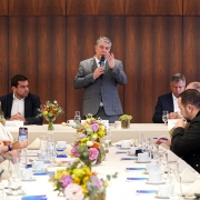 Esta é uma fotografia de um evento de negócios ou coletiva de imprensa, com um homem em pé discursando para um grupo de pessoas sentadas em uma mesa de conferência.

O homem, com cabelo grisalho e barba, veste um terno cinza e está em pé na cabeceira de uma longa mesa branca. Ele segura um microfone com a mão esquerda e gesticula com a direita enquanto fala.

A mesa está coberta por uma toalha branca e tem arranjos florais em vasos de vidro, copos de água, xícaras de café e papéis de anotação. Várias pessoas, em sua maioria homens de terno, estão sentadas ao redor da mesa, prestando atenção ao orador.

O fundo da sala é revestido com painéis de madeira de cor escura, e a iluminação é brilhante, vinda do teto.