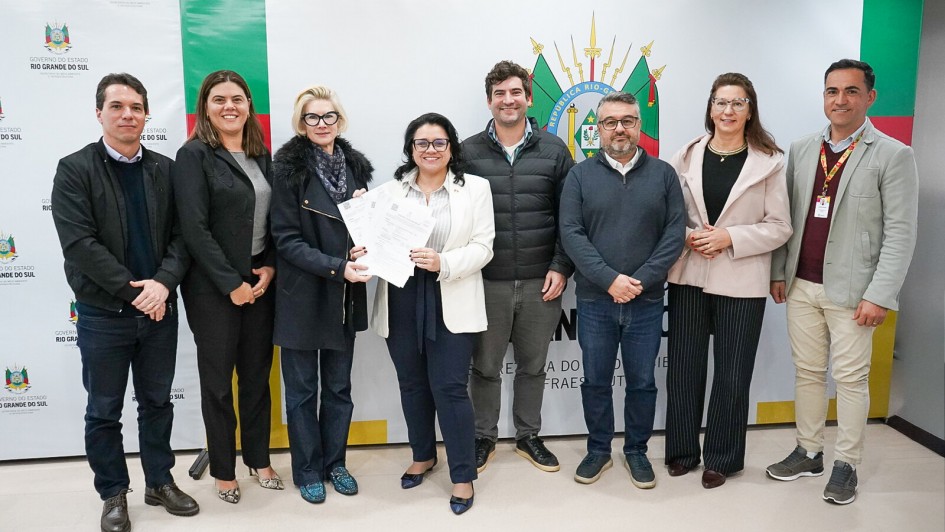 Esta é uma fotografia de um grupo de oito pessoas, quatro homens e quatro mulheres, em pé lado a lado, posando para a câmera.

No centro do grupo, uma mulher de cabelo escuro, com um blazer branco e calça azul-marinho, segura um documento com as duas mãos e sorri. Ao seu lado, à esquerda, uma mulher de cabelo loiro, usando um casaco de lã preto, também sorri. À direita da mulher central, um homem de barba com um suéter e calça jeans está posicionado.

O grupo está diante de uma parede com banners: à esquerda, há um banner vertical com o logotipo e o nome "GOVERNO DO RIO GRANDE DO SUL" em letras pretas sobre fundo verde e branco. À direita, um banner maior, branco e verde, tem o brasão do Rio Grande do Sul e a inscrição "GOVERNO DO ESTADO DO RIO GRANDE DO SUL".

Todos os participantes estão sorrindo. O ambiente é formal e bem iluminado.
