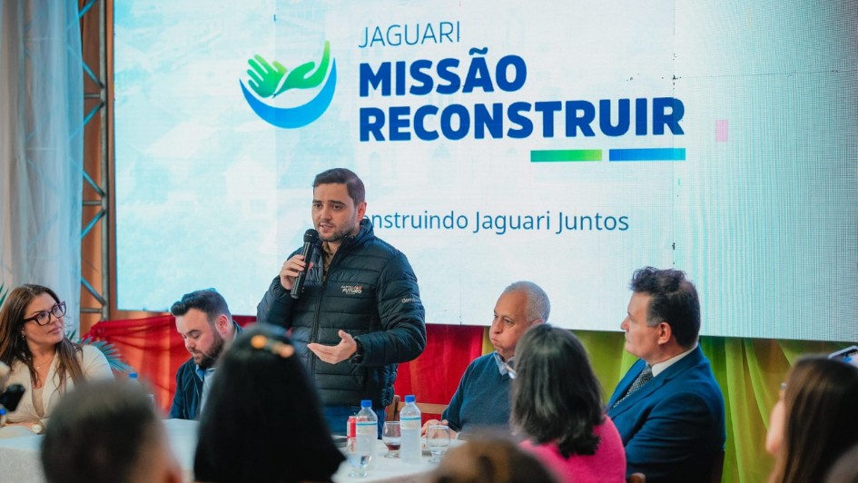 Grupo de pessoas sentadas à mesa; uma pessoa em pé fala ao microfone. Ao fundo, tela exibe “Jaguari Missão Reconstruir” e “Construindo Jaguari Juntos”.