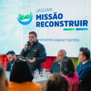 Grupo de pessoas sentadas à mesa; uma pessoa em pé fala ao microfone. Ao fundo, tela exibe “Jaguari Missão Reconstruir” e “Construindo Jaguari Juntos”.