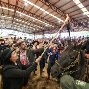 Pessoa a cavalo segura uma tocha enquanto outra encosta um bastão nela, em arena coberta. Público observa; parece cerimônia tradicional.