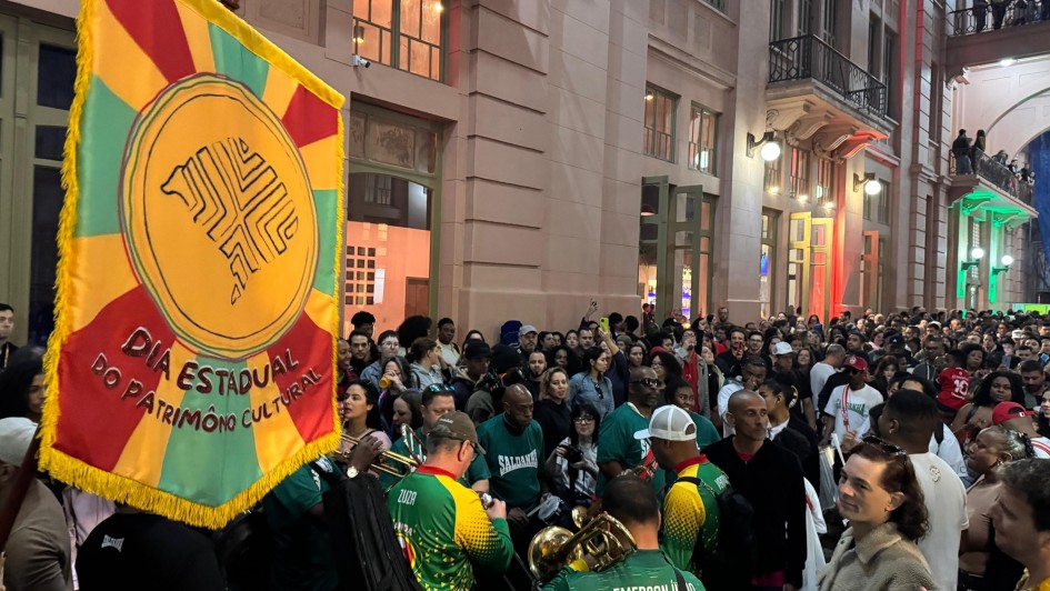 A imagem mostra um evento noturno em uma rua movimentada. Um grande estandarte, nas cores verde, amarelo e vermelho, domina o primeiro plano à esquerda. Nele está escrito "DIA ESTADUAL DO PATRIMÔNIO CULTURAL". No centro, um grupo de músicos, vestindo camisetas verdes com detalhes em amarelo, tocam instrumentos de sopro. Eles são o centro de uma multidão de pessoas, que preenche a rua. Ao fundo, à direita, há um prédio com janelas iluminadas e varandas onde mais pessoas observam o evento. O ambiente é festivo e vibrante. A foto foi tirada por Carlos Hammes, da Ascom Sedac.