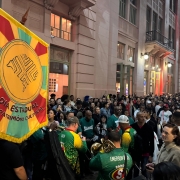 A imagem mostra um evento noturno em uma rua movimentada. Um grande estandarte, nas cores verde, amarelo e vermelho, domina o primeiro plano à esquerda. Nele está escrito "DIA ESTADUAL DO PATRIMÔNIO CULTURAL". No centro, um grupo de músicos, vestindo camisetas verdes com detalhes em amarelo, tocam instrumentos de sopro. Eles são o centro de uma multidão de pessoas, que preenche a rua. Ao fundo, à direita, há um prédio com janelas iluminadas e varandas onde mais pessoas observam o evento. O ambiente é festivo e vibrante. A foto foi tirada por Carlos Hammes, da Ascom Sedac.