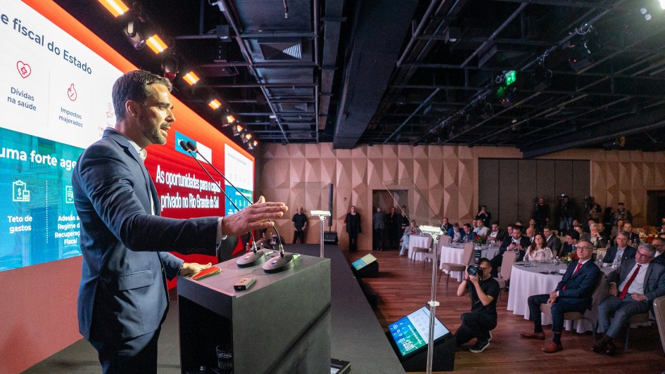Homem de terno azul escuro fala ao microfone em um palco durante um evento formal, gesticulando com uma das mãos. Ao fundo, há um grande telão colorido com gráficos e textos sobre oportunidades de investimento no Rio Grande do Sul. À direita da imagem, diversas pessoas, vestidas socialmente, estão sentadas em mesas redondas assistindo à apresentação. O ambiente é fechado, com iluminação de auditório e estrutura moderna.
