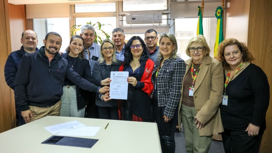 A imagem mostra um grupo de 11 pessoas, em pé, em uma sala de escritório. No centro, uma mulher de blusa preta e um xale vermelho segura um documento em suas mãos, cercada por outras pessoas que também tocam o papel. Os demais indivíduos estão em pé, sorrindo, ao redor. O ambiente tem paredes de madeira e uma grande janela ao fundo. A sala parece ser um escritório do governo, com uma bandeira do Rio Grande do Sul e uma bandeira do Brasil visíveis no canto superior direito.