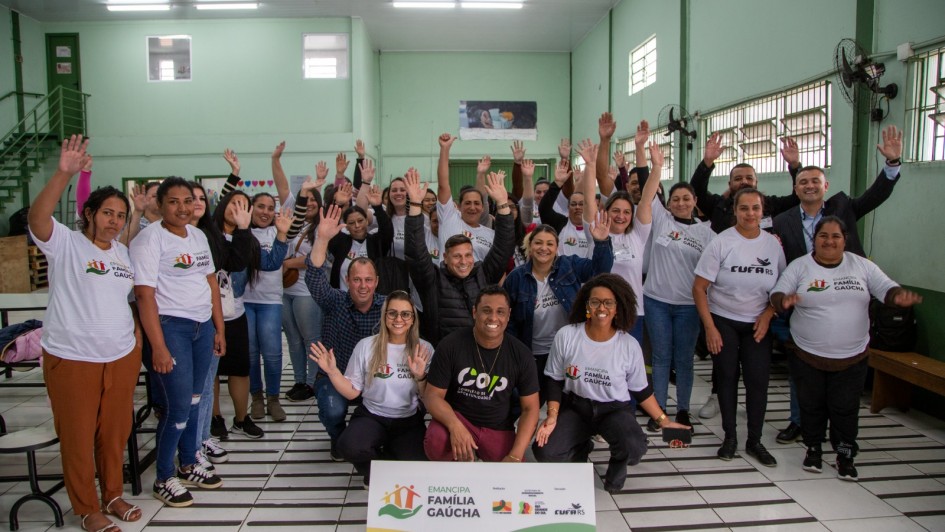 A imagem mostra um grande grupo de pessoas, de pé e agachadas, dentro de um auditório ou sala de aula. Eles estão posando para a câmera, com muitos acenando ou levantando os braços em sinal de celebração. A maioria das pessoas veste camisetas brancas com o texto "EMANCIPA FAMÍLIA GAÚCHA" e o logo da CUFA (Central Única das Favelas). Um homem no centro, agachado, veste uma camiseta preta com a palavra "CUFA". À frente do grupo, no chão, há um banner com o logo "EMANCIPA FAMÍLIA GAÚCHA" e o texto "Projeto Emancipa Família Gaúcha" e o logo da CUFA. A sala tem paredes verdes claras e um chão com ladrilhos brancos e cinza em xadrez. A atmosfera é de união e entusiasmo.