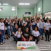 A imagem mostra um grande grupo de pessoas, de pé e agachadas, dentro de um auditório ou sala de aula. Eles estão posando para a câmera, com muitos acenando ou levantando os braços em sinal de celebração. A maioria das pessoas veste camisetas brancas com o texto "EMANCIPA FAMÍLIA GAÚCHA" e o logo da CUFA (Central Única das Favelas). Um homem no centro, agachado, veste uma camiseta preta com a palavra "CUFA". À frente do grupo, no chão, há um banner com o logo "EMANCIPA FAMÍLIA GAÚCHA" e o texto "Projeto Emancipa Família Gaúcha" e o logo da CUFA. A sala tem paredes verdes claras e um chão com ladrilhos brancos e cinza em xadrez. A atmosfera é de união e entusiasmo.