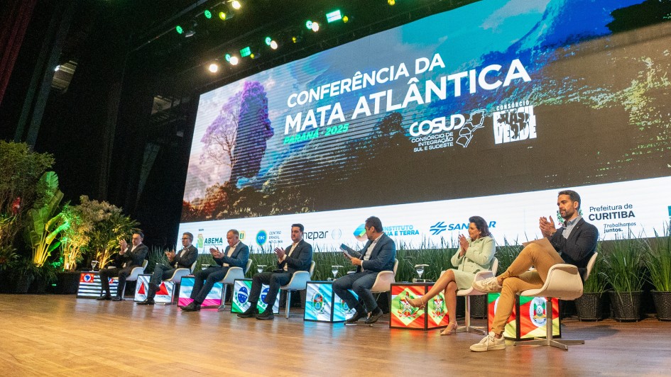 A fotografia mostra um grupo de sete pessoas sentadas em cadeiras brancas em um palco, aplaudindo durante um evento. Eles são governadores e representantes dos estados do Sul e Sudeste. Ao lado direito, em destaque, está o governador Eduardo Leite, que veste um blazer escuro e calças bege e está aplaudindo. Todos estão voltados para o centro do palco. Ao fundo, um telão de LED exibe o título "CONFERÊNCIA DA MATA ATLÂNTICA" e o logo do "COSUD", com a data e cidade "CURITIBA 2025". O palco é decorado com plantas verdes.