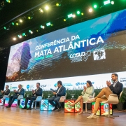 A fotografia mostra um grupo de sete pessoas sentadas em cadeiras brancas em um palco, aplaudindo durante um evento. Eles são governadores e representantes dos estados do Sul e Sudeste. Ao lado direito, em destaque, está o governador Eduardo Leite, que veste um blazer escuro e calças bege e está aplaudindo. Todos estão voltados para o centro do palco. Ao fundo, um telão de LED exibe o título "CONFERÊNCIA DA MATA ATLÂNTICA" e o logo do "COSUD", com a data e cidade "CURITIBA 2025". O palco é decorado com plantas verdes.